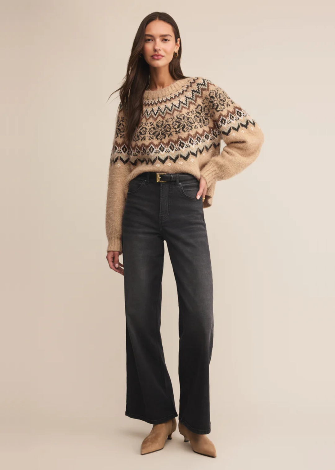 Woman wearing a brown fair isle sweater with black jeans against a tan background
