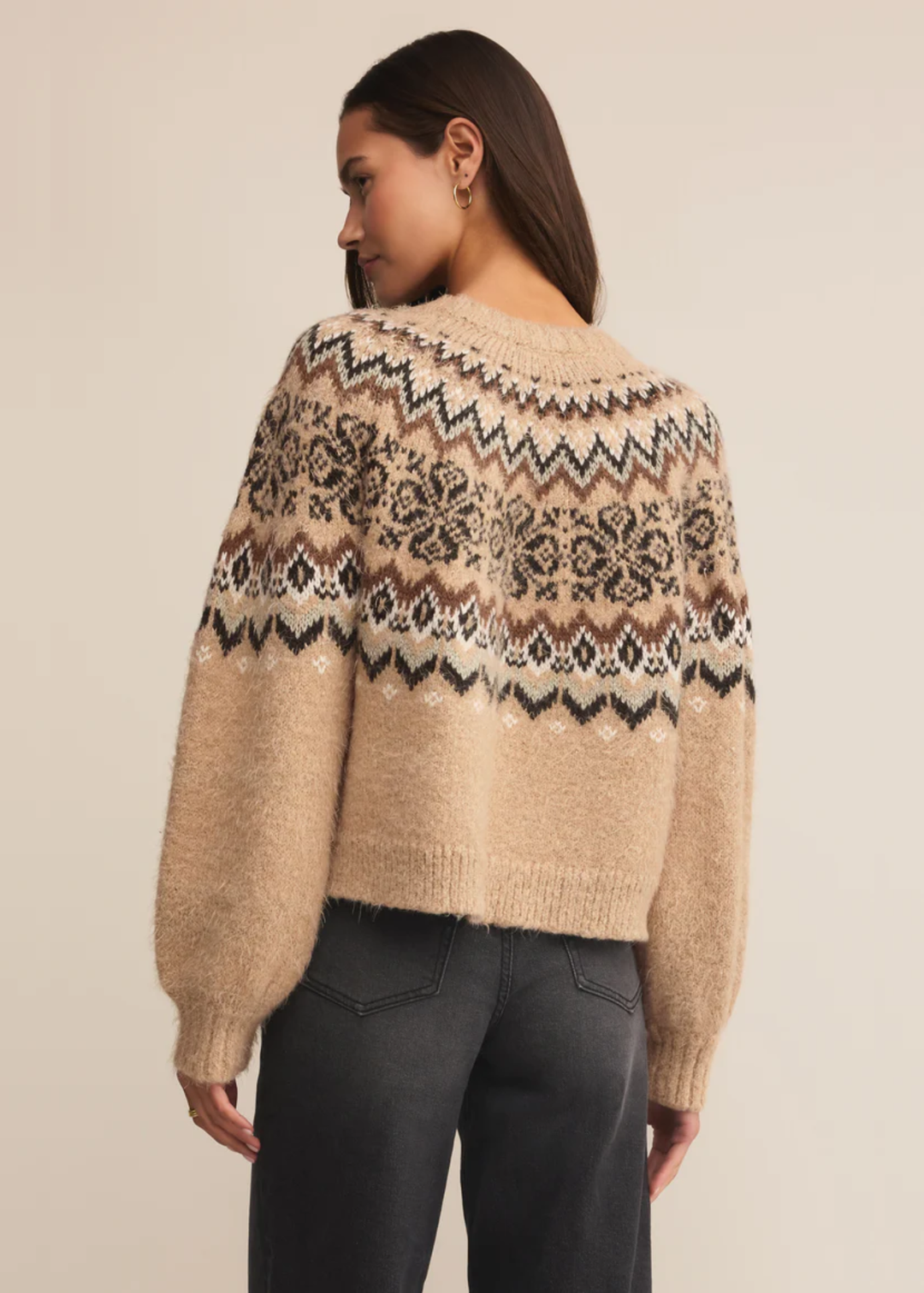 Woman turned around wearing a brown fair isle sweater with black jeans against a tan background