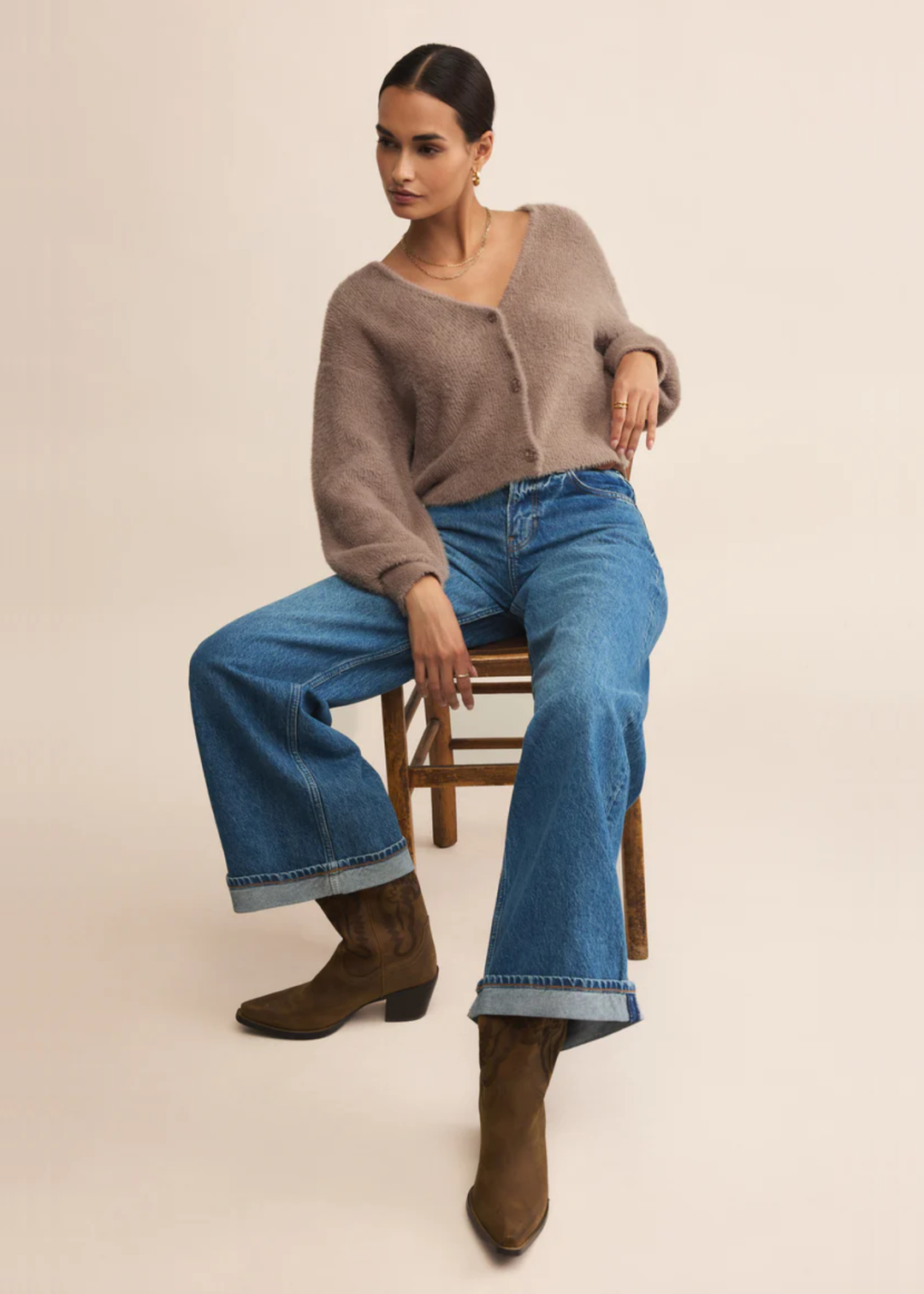 Woman sitting in a chair wearing a fuzzy light brown cardigan with jeans in front of a grey background