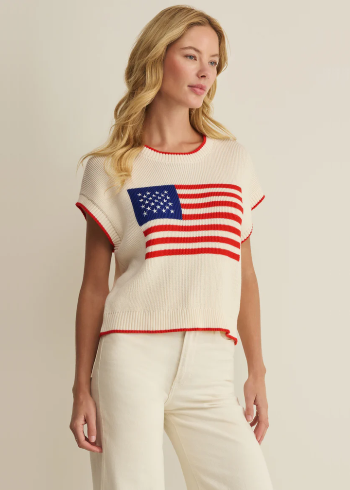 Woman wearing a cream sleeveless sweater with an American flag graphic and white jeans