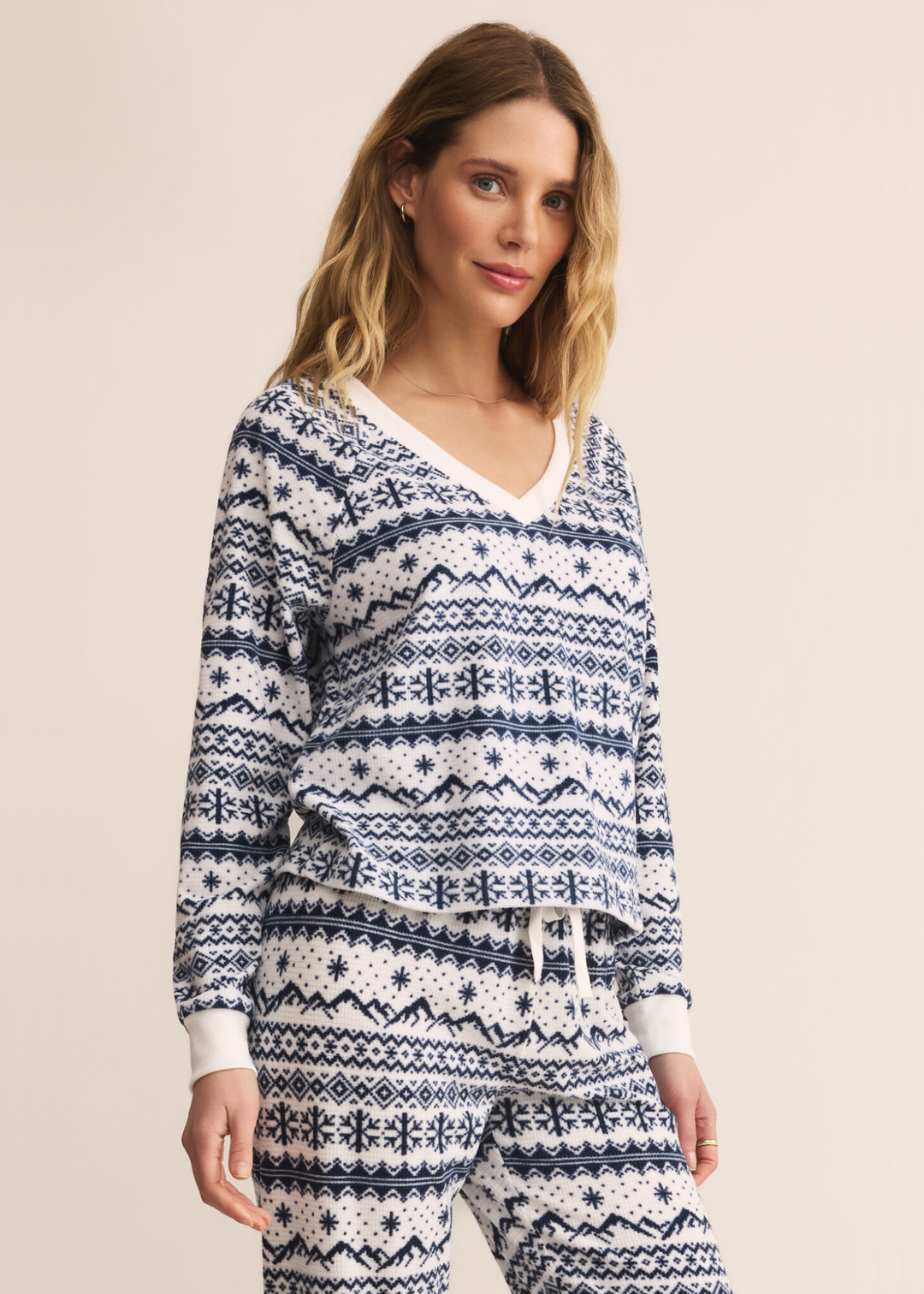 Woman turned to the side wearing a blue and white fairisle patterned v-neck pajama top with matching pants in front of a tan background