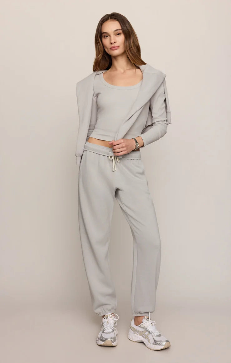 A woman modeling a light gray two-piece lounge set consisting of a cropped tank top with a draped cardigan-style layer and matching jogger pants, paired with white sneakers, against a solid light beige studio background.