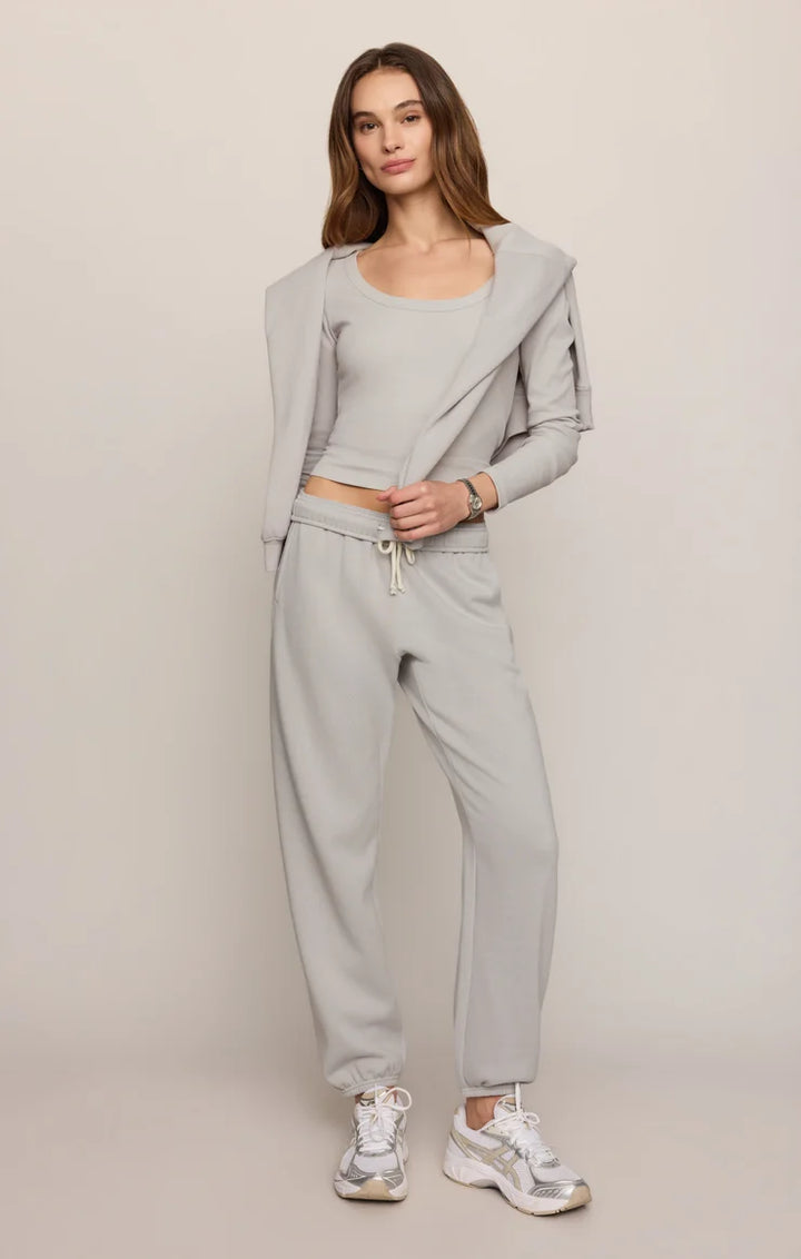 A woman modeling a light gray two-piece lounge set consisting of a cropped tank top with a draped cardigan-style layer and matching jogger pants, paired with white sneakers, against a solid light beige studio background.