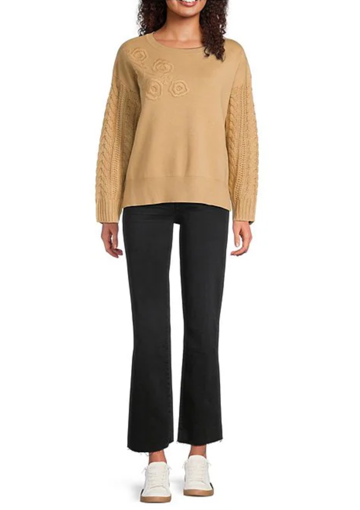 A woman modeling a tan crewneck sweater with textured cable-knit sleeves and circular knit details on the chest, paired with black cropped flare pants and white sneakers against a plain white background.