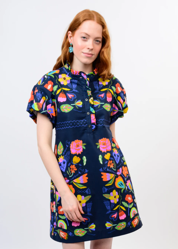 A woman modeling a short-sleeve navy blue dress with a vibrant multicolor floral and butterfly print, set against a plain light gray background.