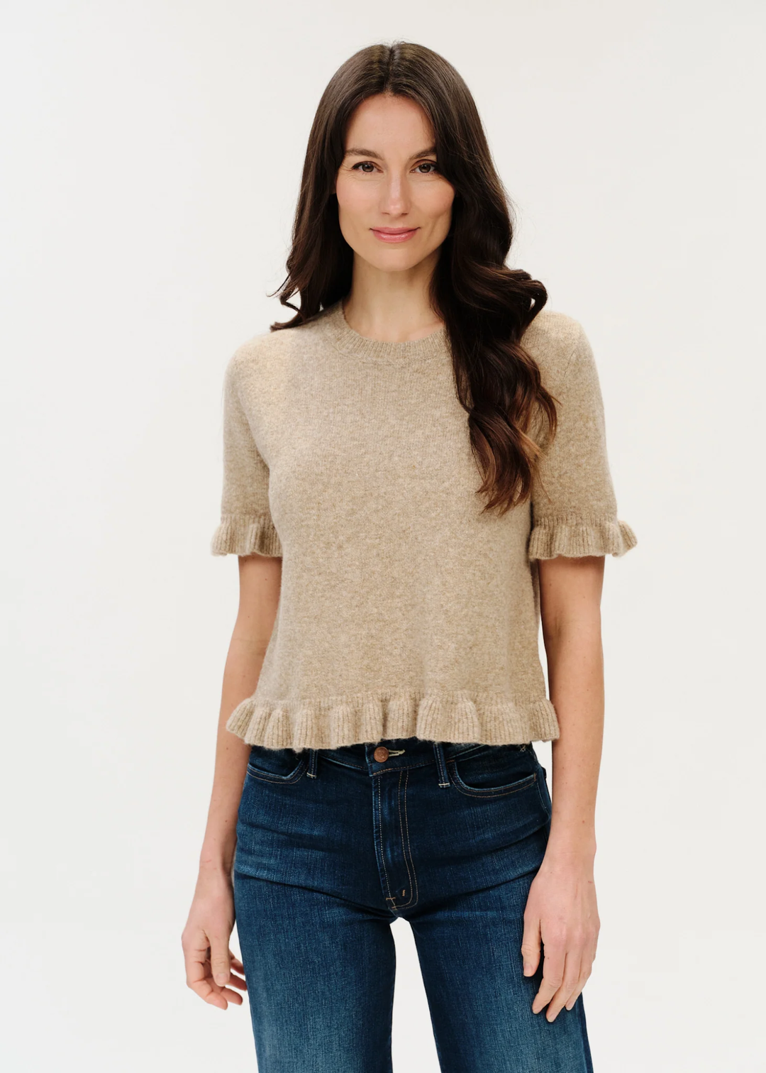 A woman modeling a short-sleeve knit top in a light beige color with ruffled cuffs and a ruffled hem, paired with dark blue jeans, against a plain white background.