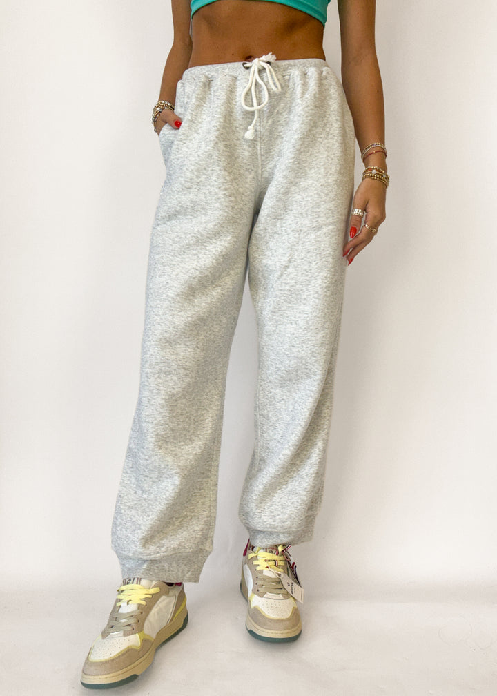 A person wearing light heather gray jogger sweatpants with a white drawstring, paired with beige sneakers, shown from the waist down against a plain light background.