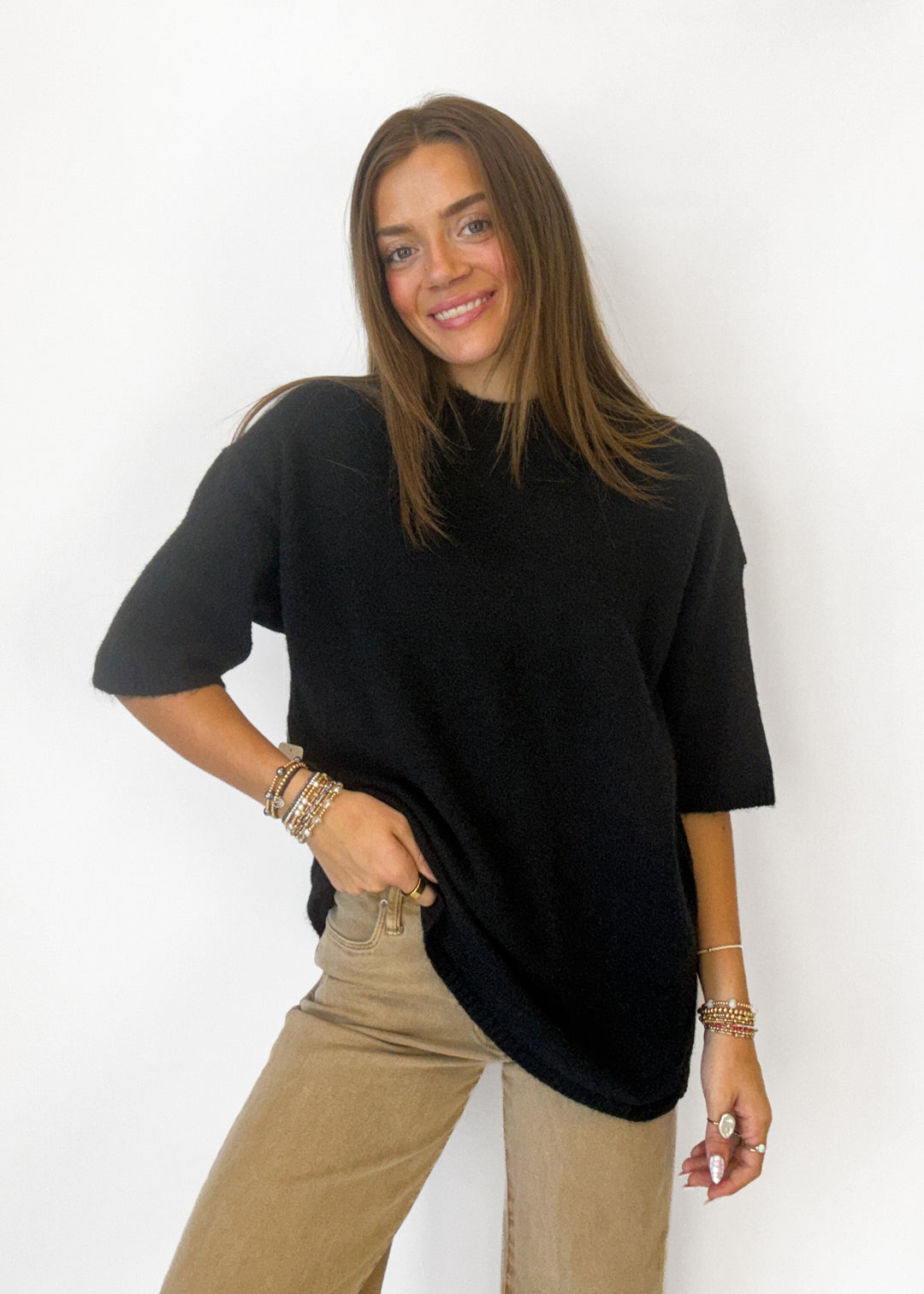 A woman modeling a black short-sleeve knit top with a high mock neck, paired with beige pants, against a plain white background.