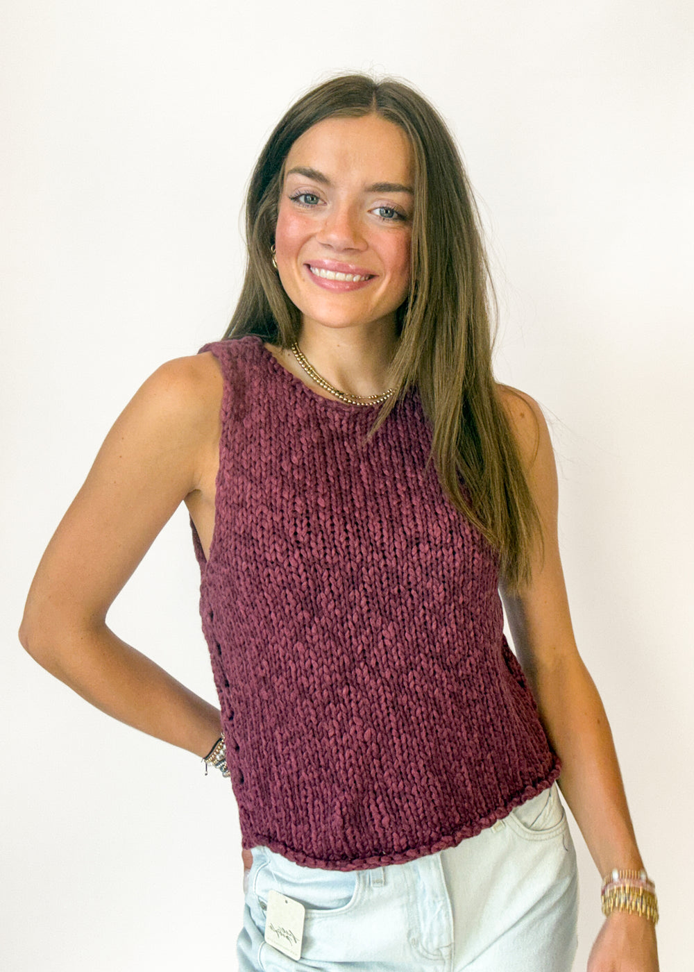 A smiling young woman with long light brown hair wearing a sleeveless, ribbed knit top in a heathered burgundy shade, paired with light-wash denim shorts, against a plain white background.