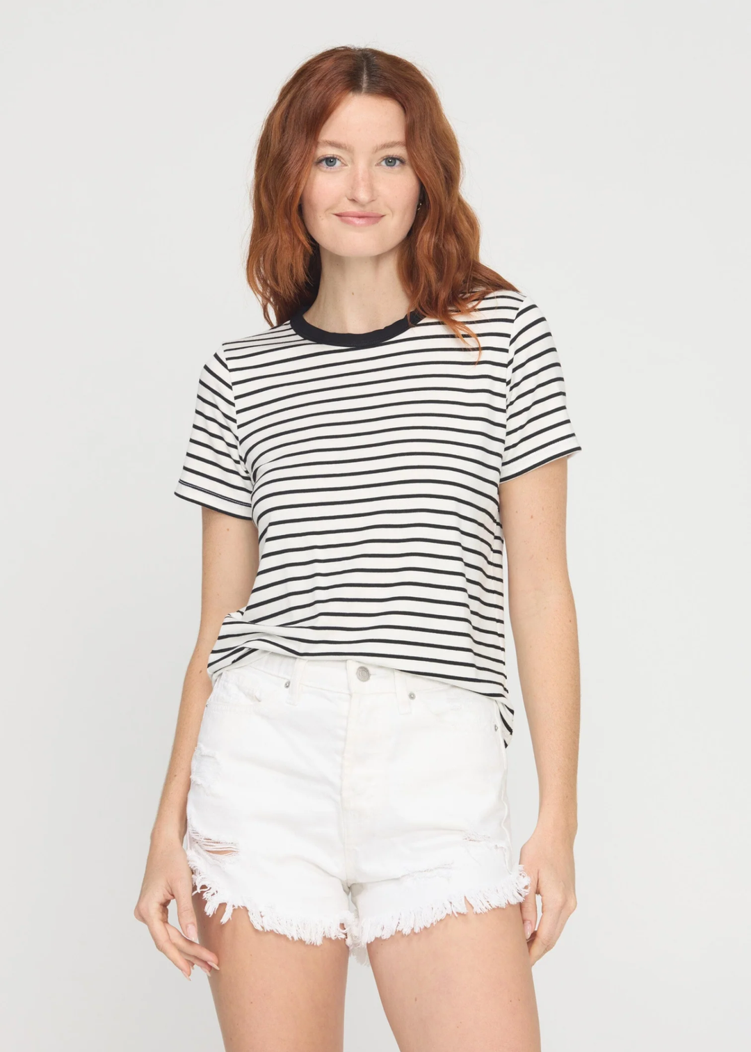 A woman with red hair wearing a black and white horizontally striped short-sleeve t-shirt with a black crew neck, paired with white denim shorts, against a light gray studio background.