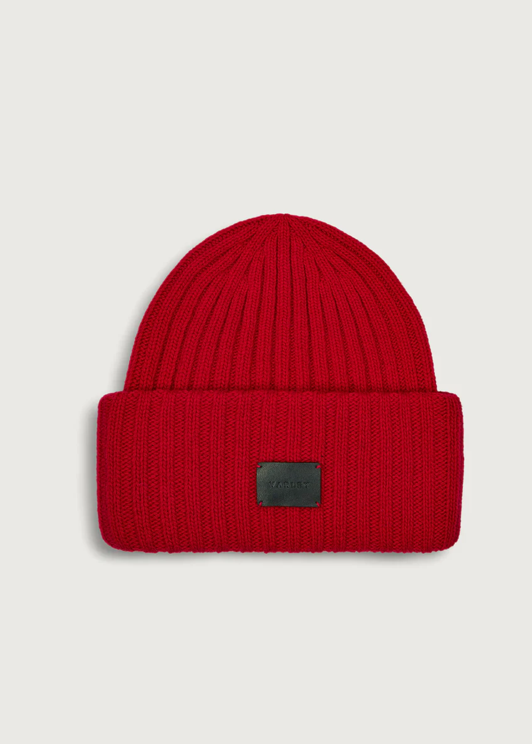 A bright red ribbed knit beanie hat with a folded cuff and a small black rectangular logo patch on the front, shown against a light gray background.