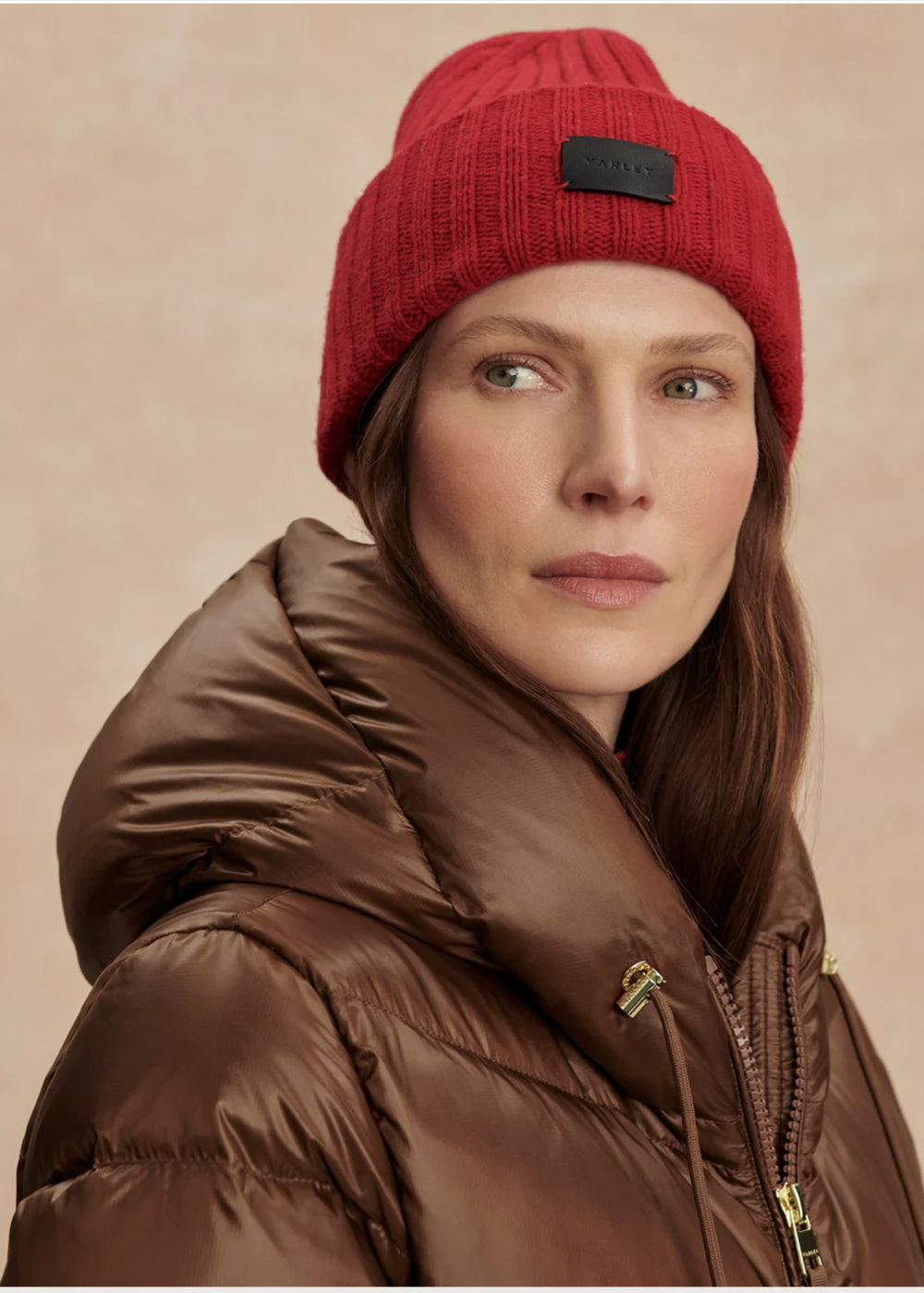 A woman wearing a red ribbed knit beanie with a black rectangular logo patch and a brown puffer jacket, photographed against a warm beige studio background.