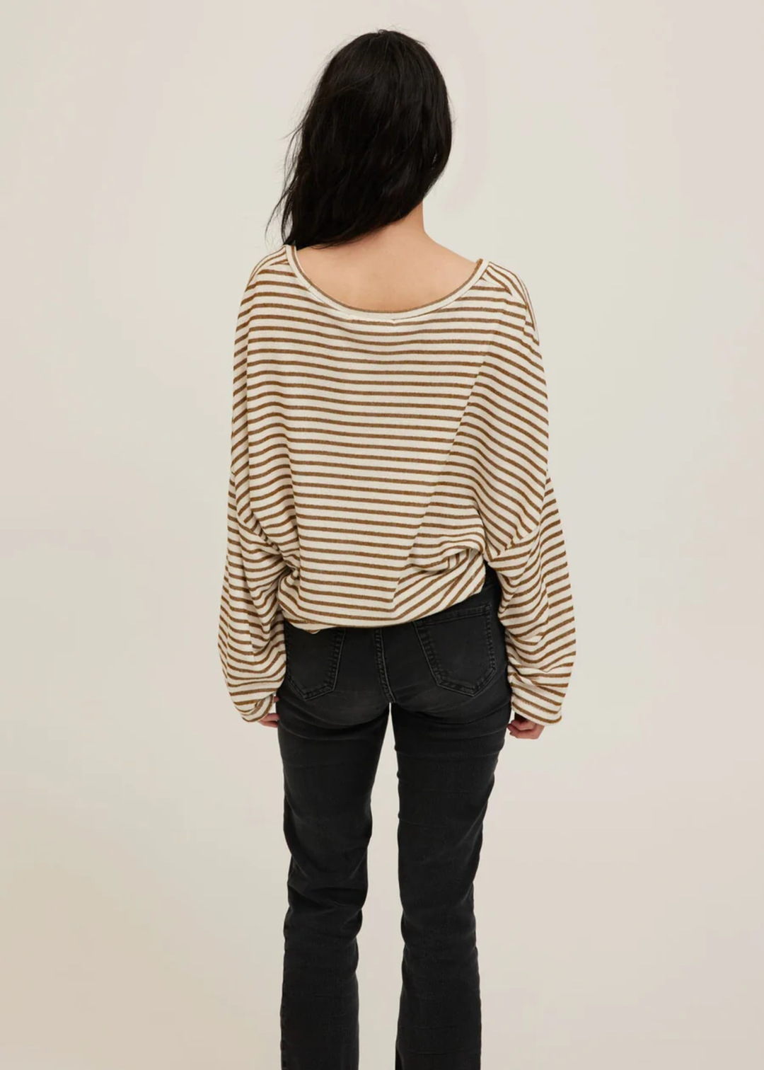 A woman viewed from behind wearing a beige and brown horizontally striped long-sleeve top and black jeans, set against a plain light beige background.