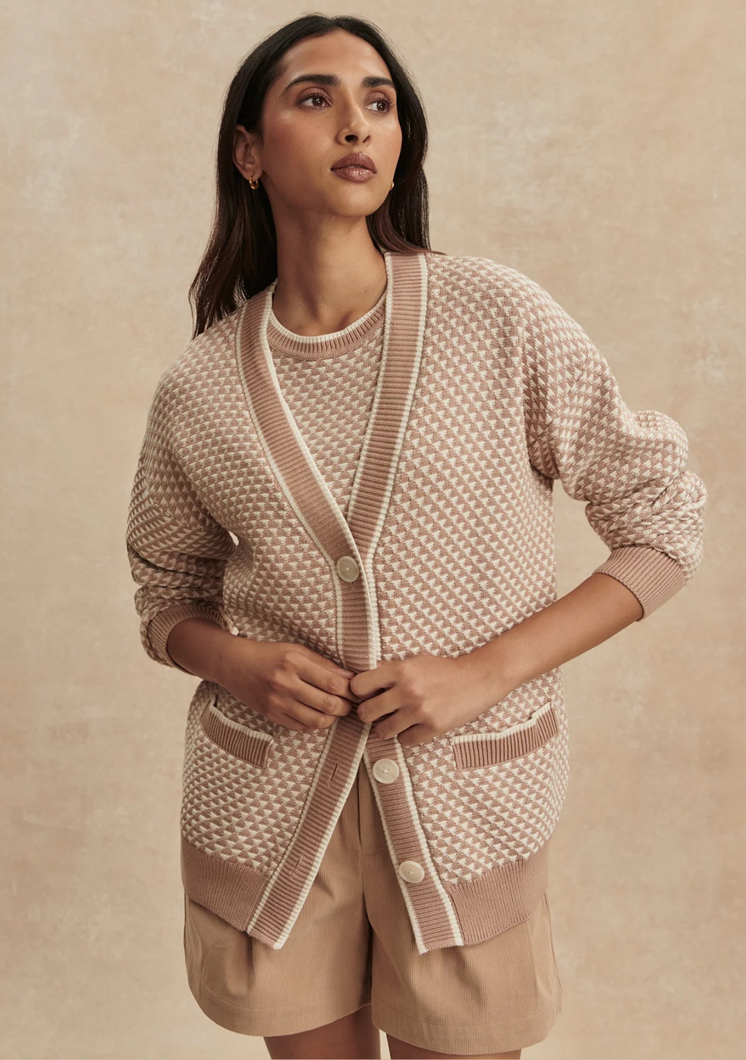 Woman wearing a beige and brown patterned cardigan over a matching top with beige shorts on a beige background.