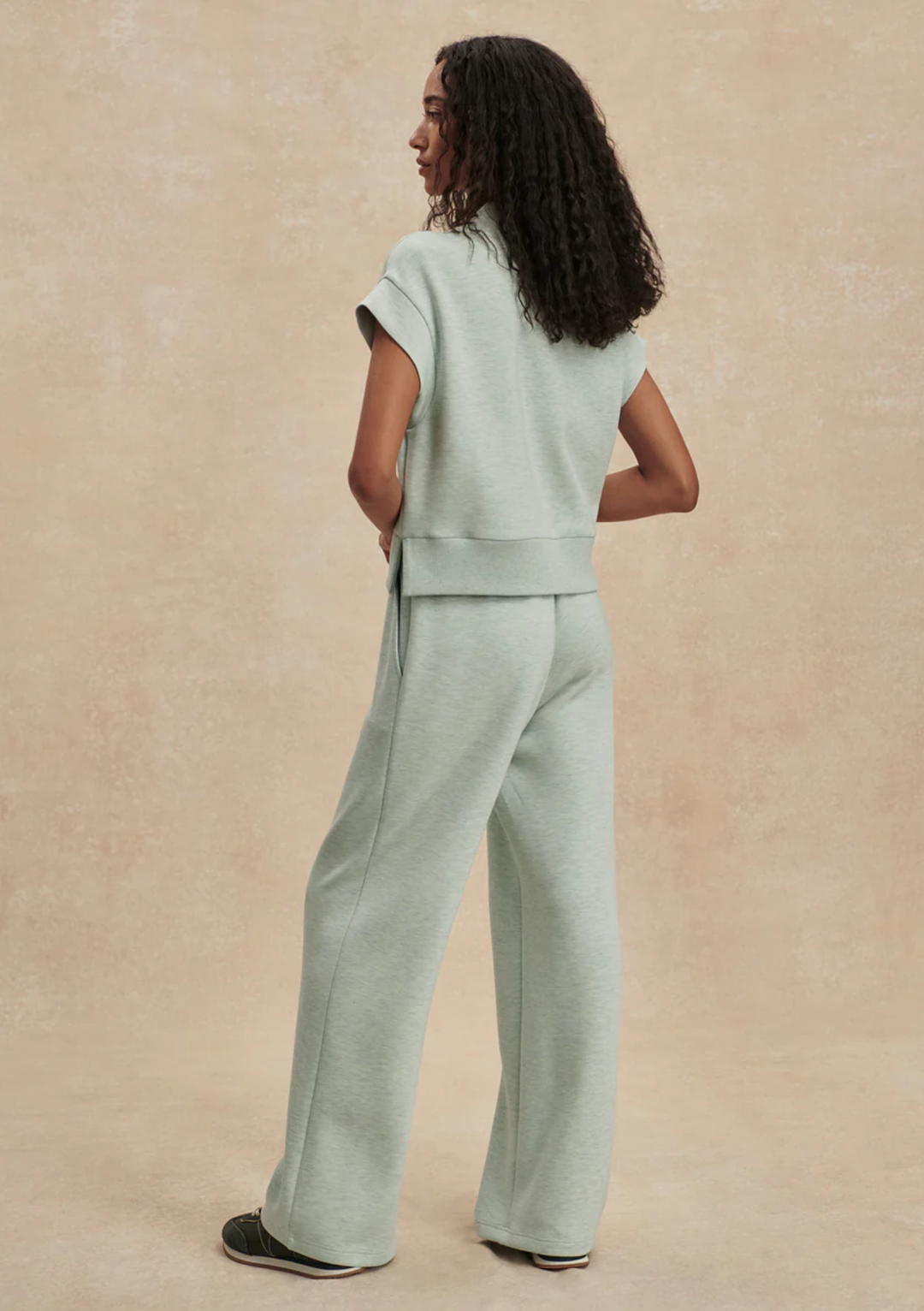 A woman wearing a light sage green two-piece lounge set, consisting of a short-sleeve cropped top and matching wide-leg pants, shown from a back-three-quarter view against a textured beige studio backdrop.