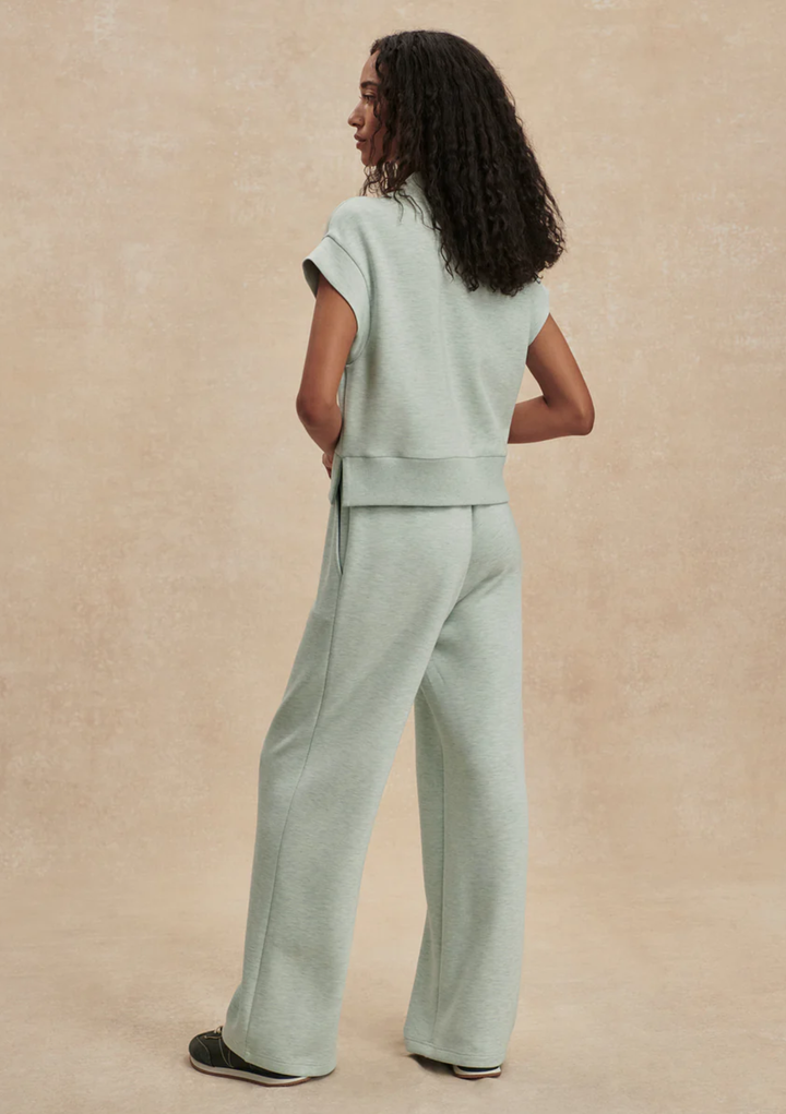 A woman wearing a light sage green two-piece lounge set, consisting of a short-sleeve cropped top and matching wide-leg pants, shown from a back-three-quarter view against a textured beige studio backdrop.