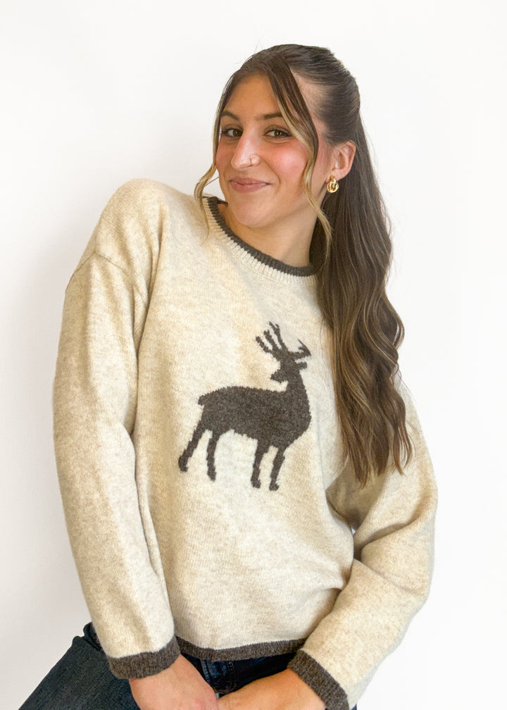A woman wearing a cream-colored knit sweater with a dark brown reindeer graphic on the front, set against a plain white background.