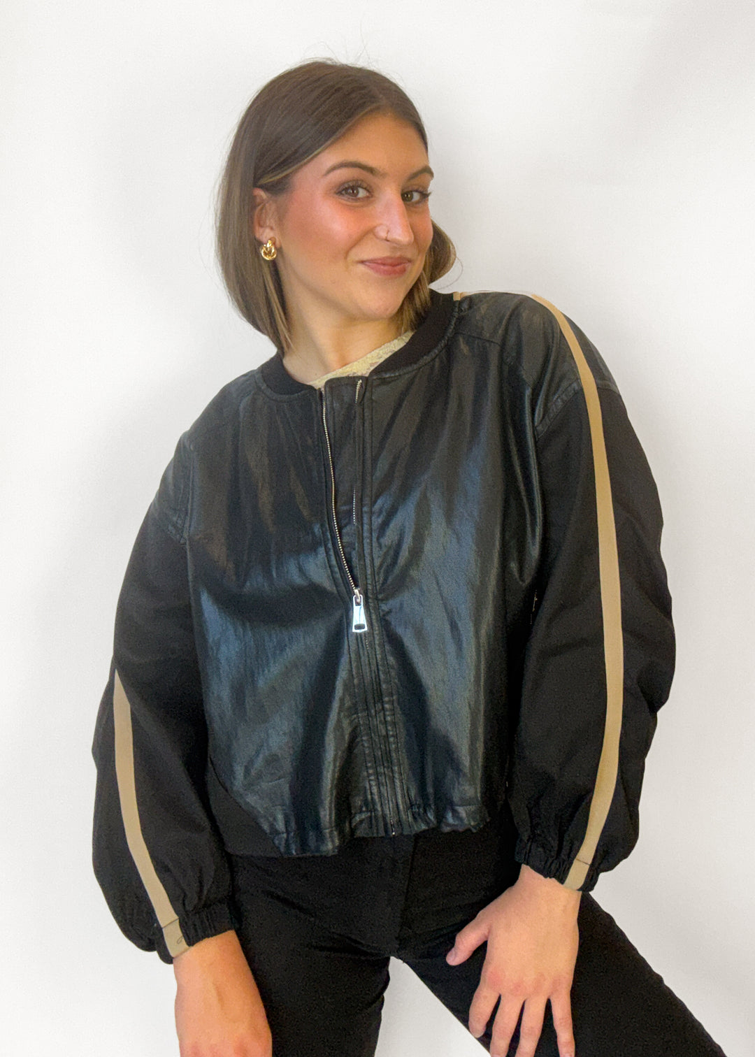 A woman wearing a shiny black bomber jacket with beige stripes on the sleeves, paired with black pants, posing against a white studio background.