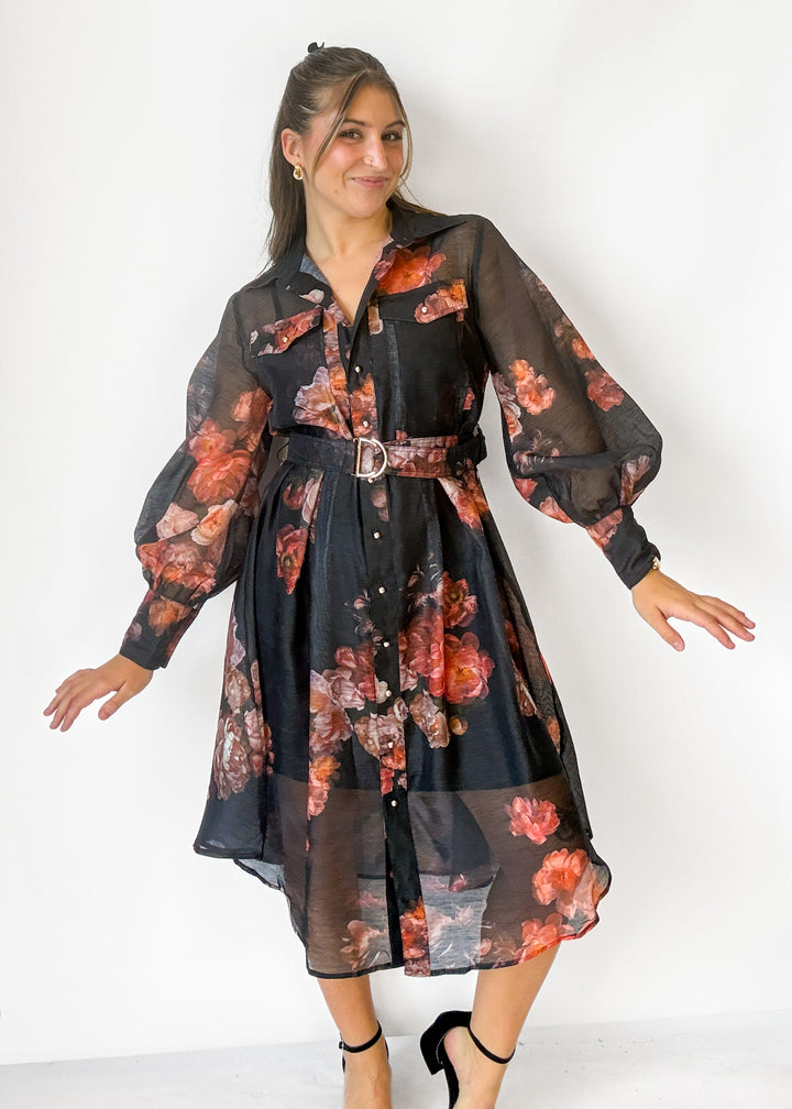 A woman modeling a sheer black midi shirt dress with a red and orange floral print, featuring balloon sleeves, a button-down front, and a matching fabric belt with a silver buckle.