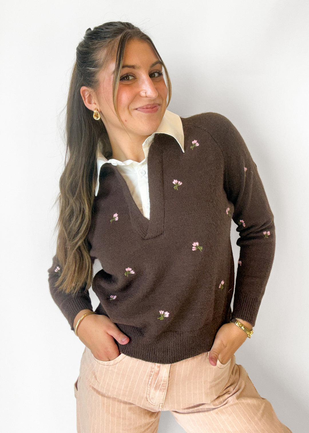 A woman wearing a dark brown knit sweater with small pink and green floral embroidery, layered over a white collared shirt and paired with light pink pinstripe trousers, set against a plain white background.
