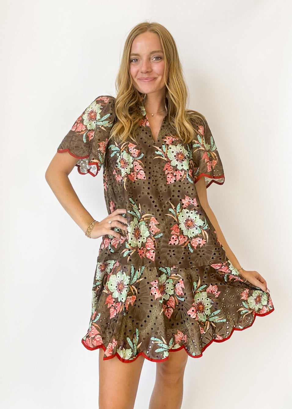 A woman modeling a brown short-sleeve mini dress with a colorful floral print, featuring red scalloped trim on the sleeves and hem, against a plain white background.