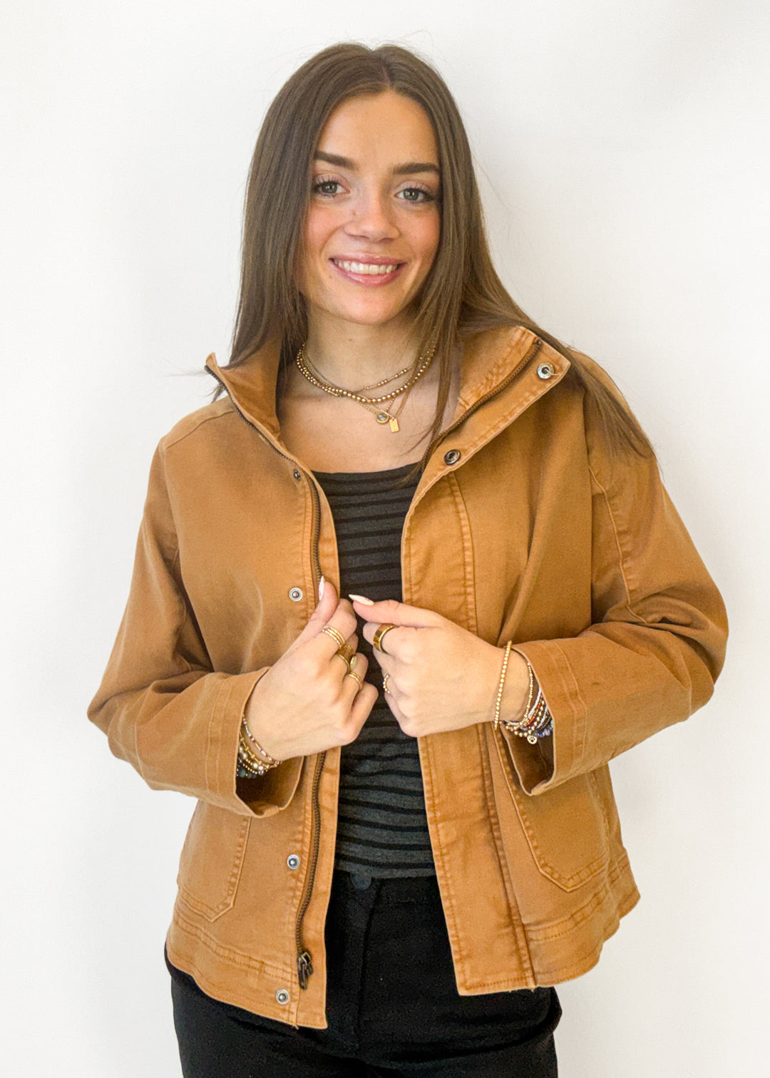 A woman wearing a cropped tan-brown utility jacket over a black striped top and black pants, standing against a plain white background.
