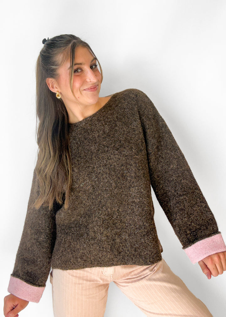 A woman wearing a brown textured knit sweater with light pink ribbed cuffs, paired with light pink pants, posing against a plain white wall.