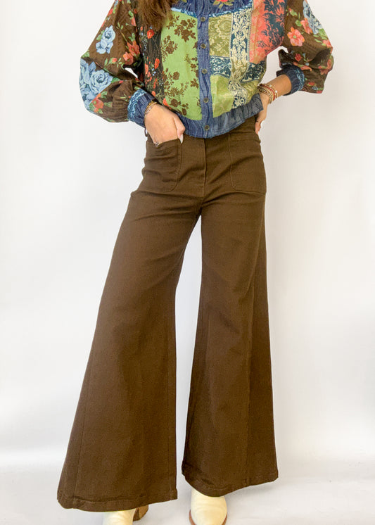 A person wearing brown wide-leg pants and a multicolored patchwork floral blouse, standing against a plain white studio background.