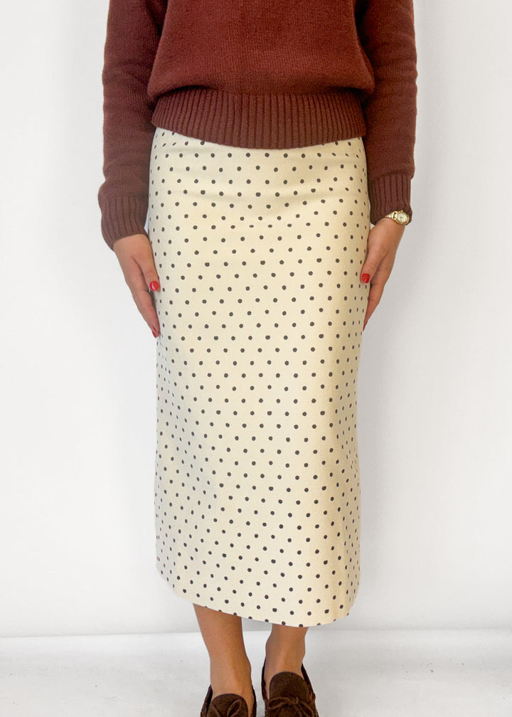 A person wearing a cream-colored midi skirt with a black polka dot pattern, paired with a rust-brown sweater and brown suede loafers, standing against a plain white wall.