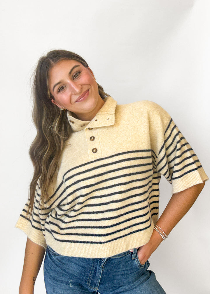 A woman wearing a cream knit polo sweater with black horizontal stripes and a three-button placket, paired with blue jeans, photographed against a plain white studio background.