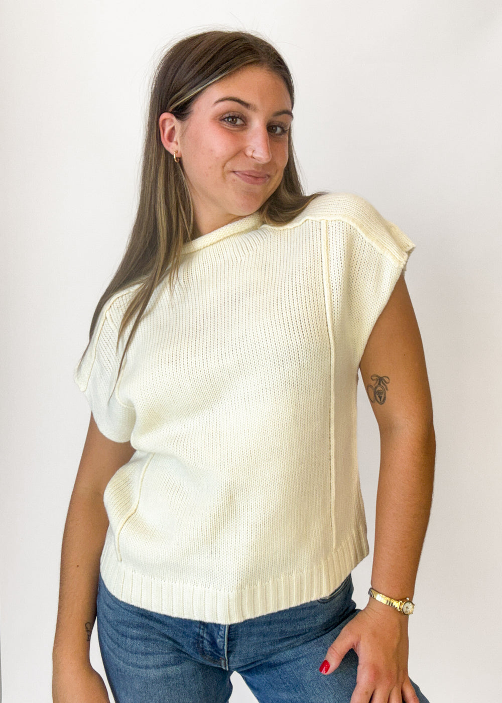 A woman wearing a cream-colored ribbed knit sleeveless sweater vest over a white top, paired with blue jeans, standing against a plain white wall.