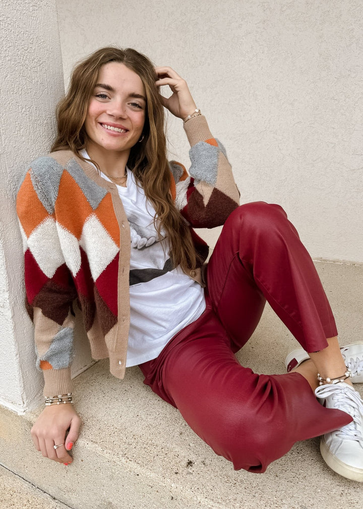 Fall Argyle Cardigan in Oatmeal Multi featuring a bold, geometric pattern with cozy textured details, styled over a white graphic tee and red coated flare pants.