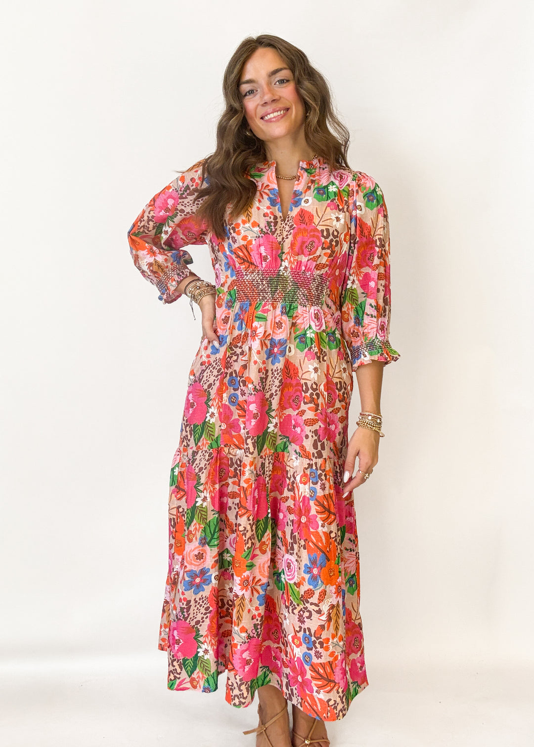 A woman modeling a long-sleeve maxi dress with a vibrant, dense floral print in pink, orange, green, and blue on a cream background, set against a plain white studio backdrop.