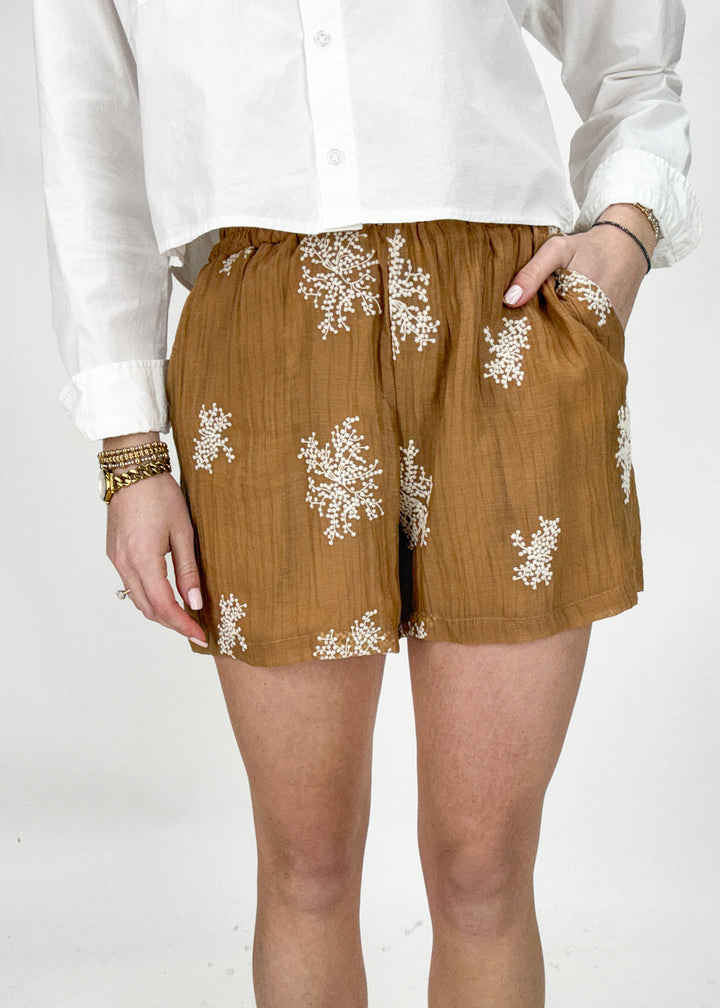 Gold shorts with white floral patterns worn by a person on a white background