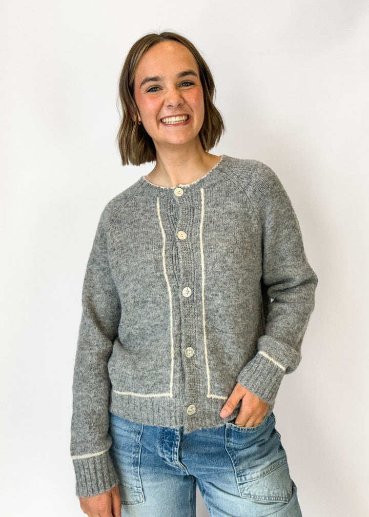 A smiling woman with short brown hair wearing a gray knit cardigan with cream-colored trim and buttons, paired with blue jeans, standing against a plain white background.