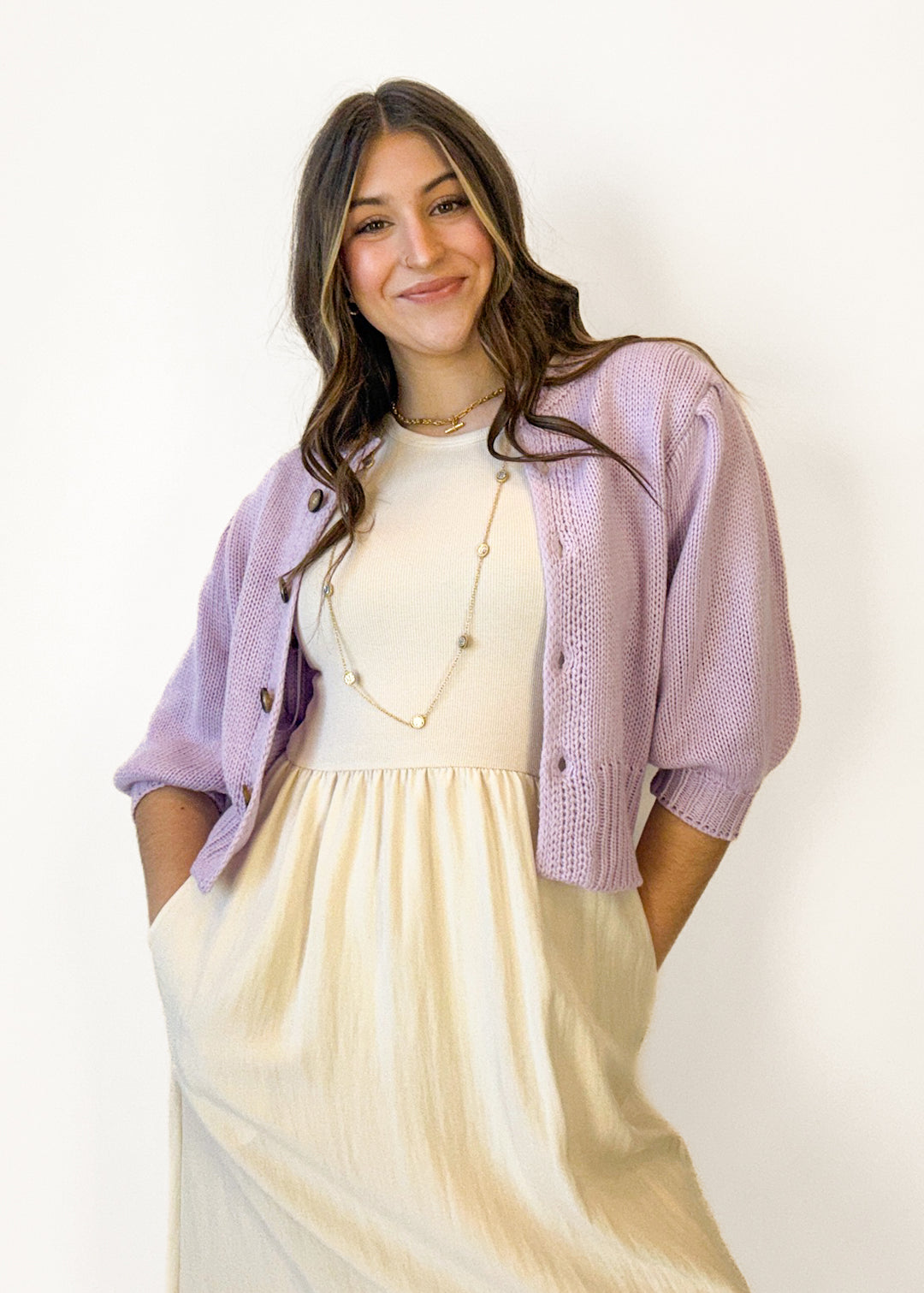 Woman wearing a light purple cardigan over a white top and beige skirt on a plain background