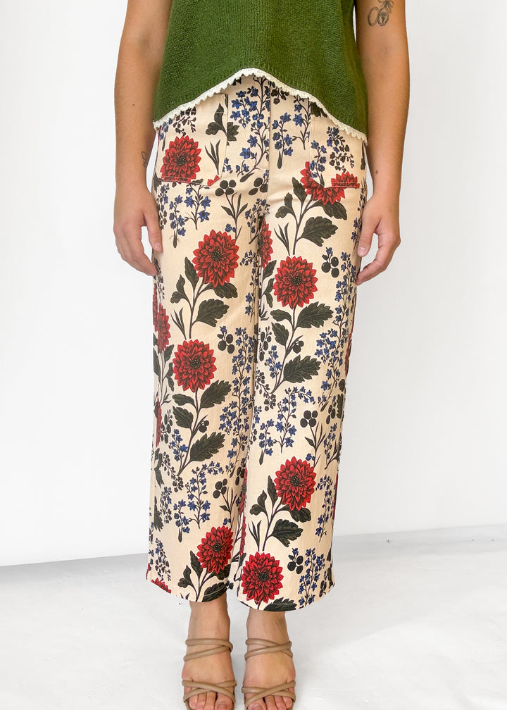 A person wearing wide-leg cropped pants with a beige base and a red, blue, and black floral print, paired with a green top and tan strappy heels against a white studio background.