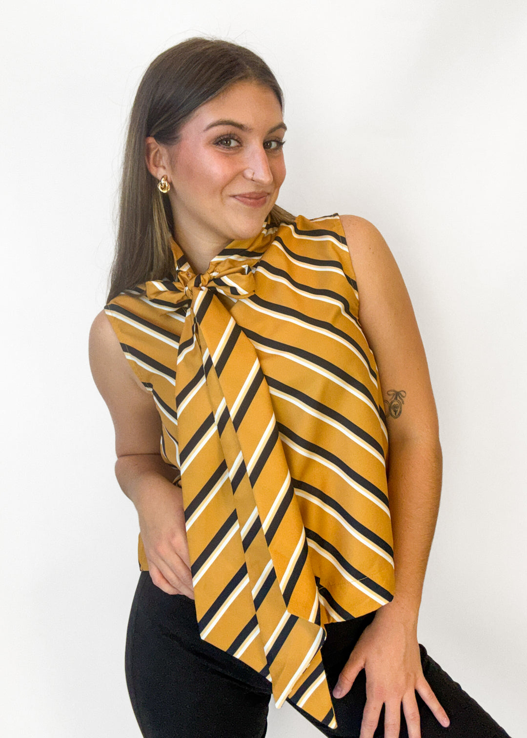 A woman modeling a sleeveless yellow top with diagonal black and white stripes and a large front bow, paired with black pants, against a plain white background.