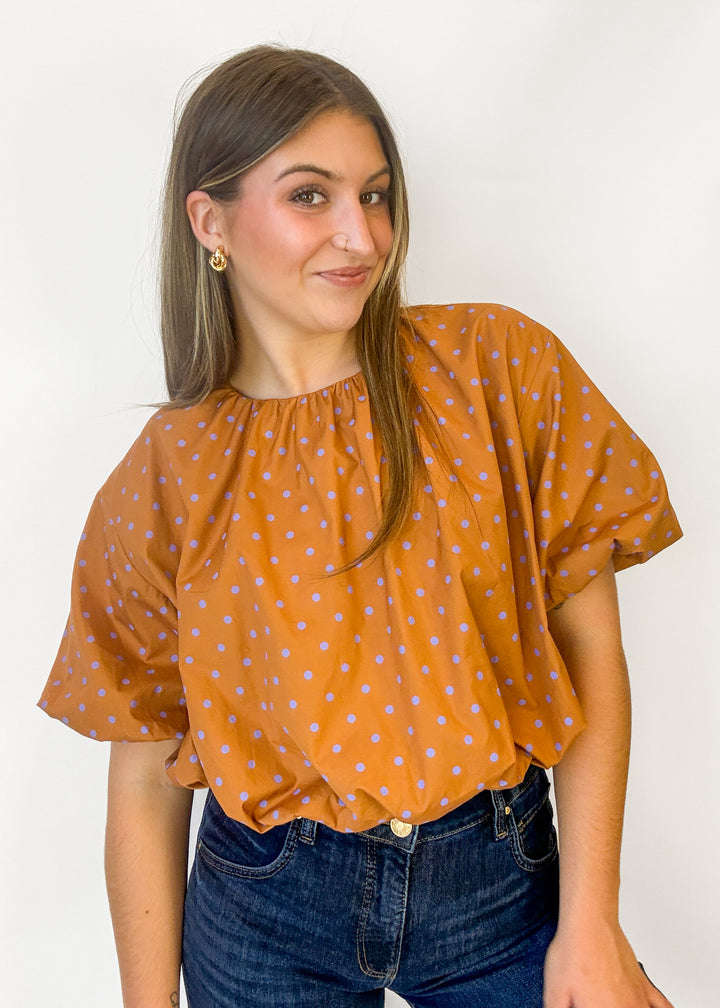 A woman wearing a mustard orange short-sleeve blouse with light blue polka dots and dark blue jeans, standing against a plain white background.