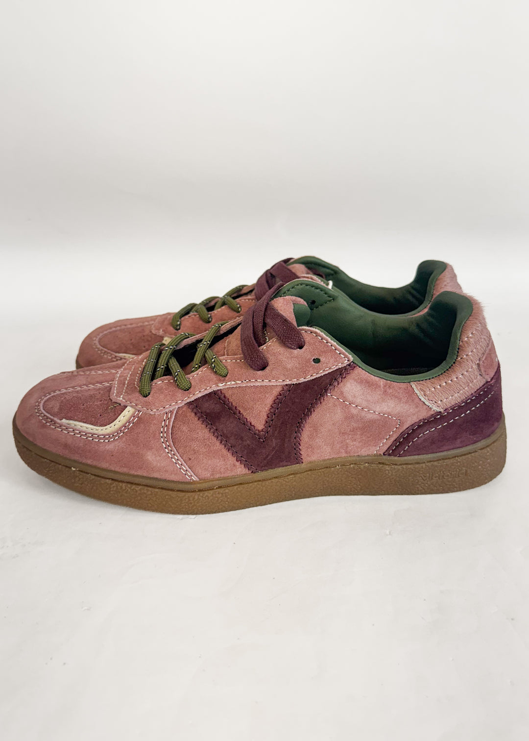 A pair of low-top suede sneakers in dusty rose pink and deep plum purple, featuring olive green laces, a green interior lining, and brown gum soles, photographed on a light gray studio background.