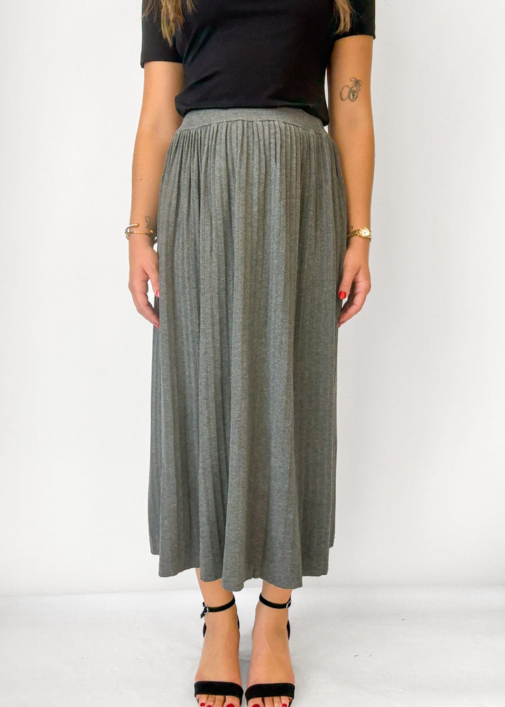 A woman wearing a high-waisted, pleated gray maxi skirt with a black short-sleeve top and black high-heeled sandals, standing against a plain white studio background.
