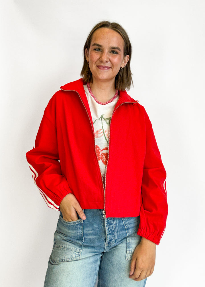 A woman wearing a bright red zip-up track jacket with white stripes on the sleeves,