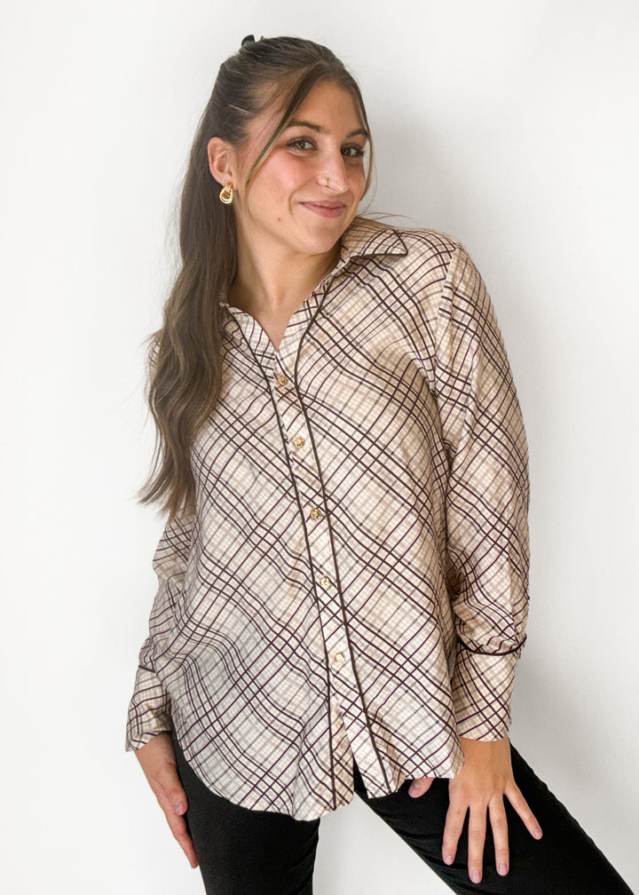 A woman wearing a beige and brown plaid button-up shirt with black pants, posing against a plain white wall.