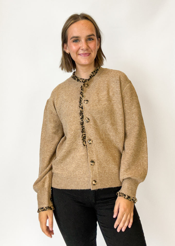 A woman modeling a beige knit cardigan with leopard-print trim along the collar, front button placket, and cuffs, paired with black pants against a plain white studio background.