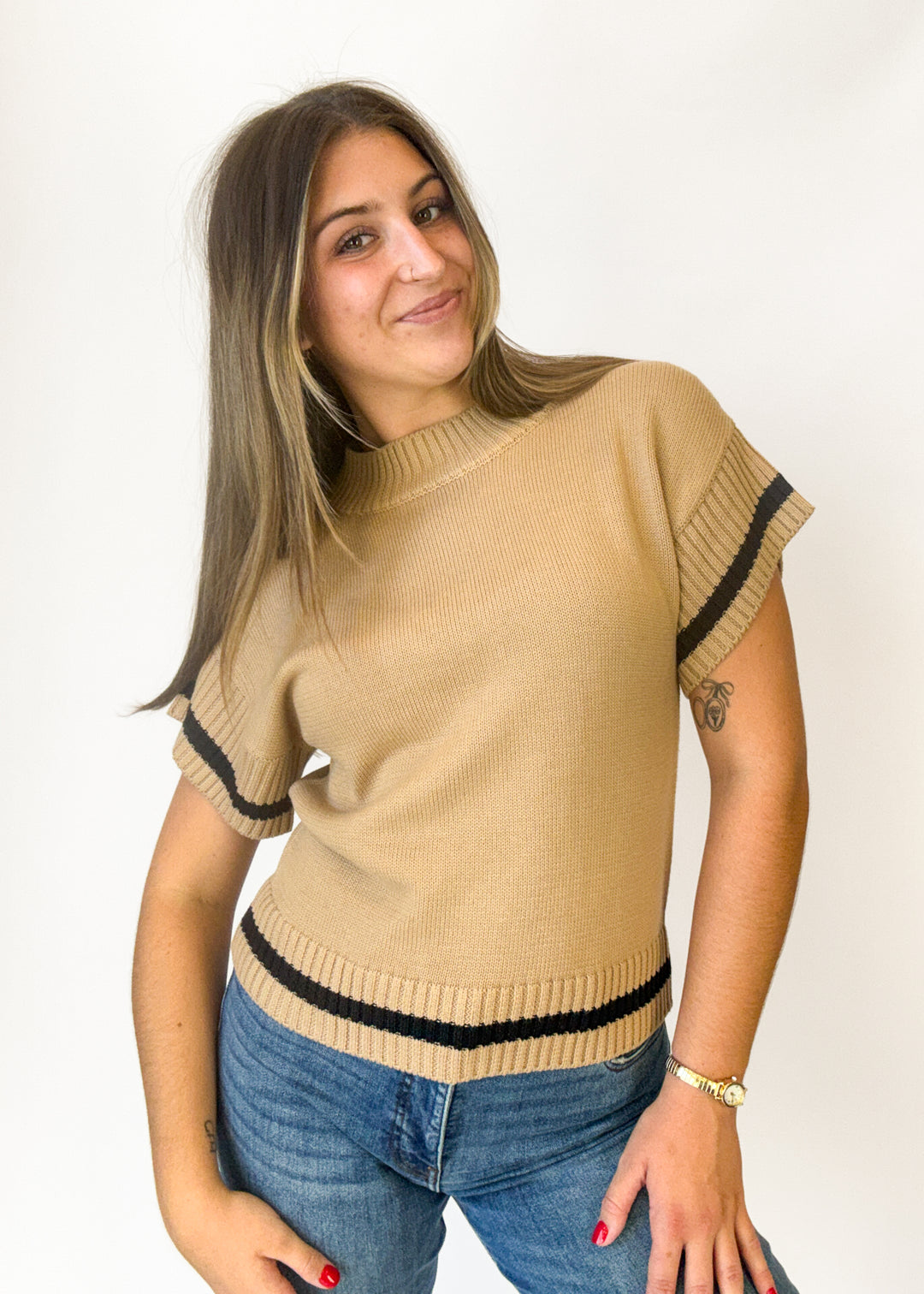 A woman modeling a beige short-sleeve knit sweater with black ribbed trim at the sleeve cuffs and hem, paired with blue jeans against a plain white background.