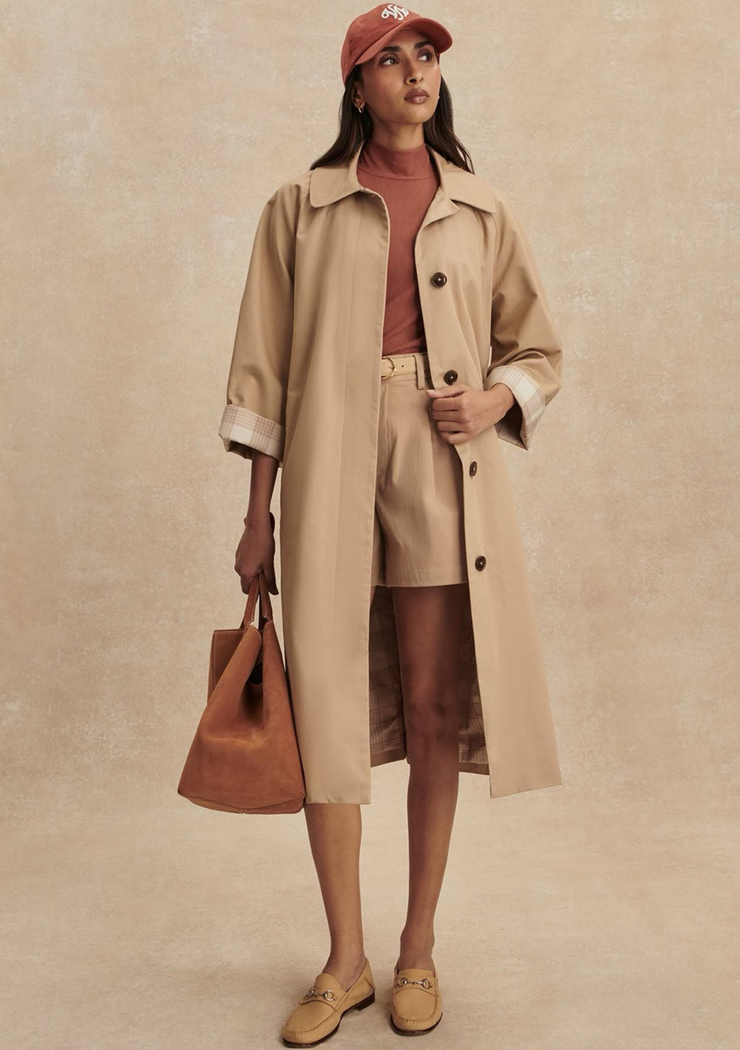 Woman wearing a beige trench coat and shorts with a brown bag against a beige background